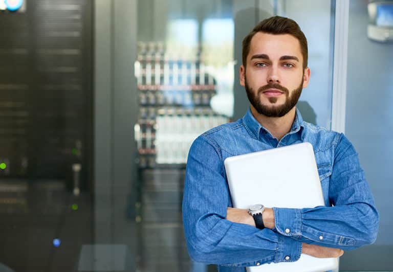 A systems administrator in a blue button-down long-sleeve shirt posing holding a laptop in front of a server cabinet and looking at camera with an air of confidence, ready to solve a technical issue thrown his way.