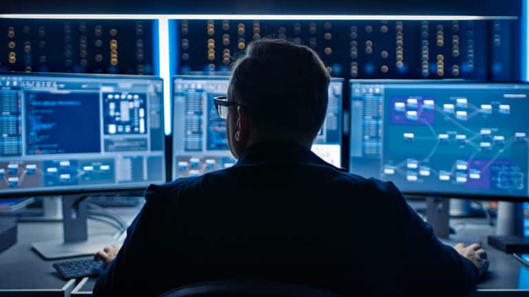 A cloud security solution specialist monitors an organization’s cloud deployments on three monitors with server racks seen in the background.