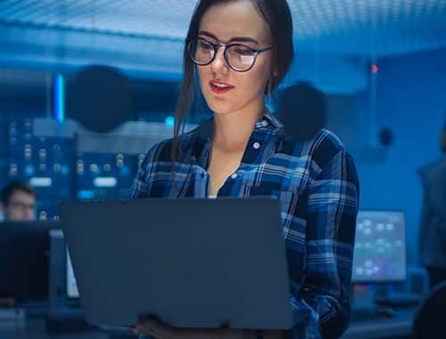 A smart attractive young woman, IT security professional wearing glasses holds a laptop while investigating a cybersecurity event. In the background of the security operations center, security specialists can be seen working on data servers racks and solving IT tickets.