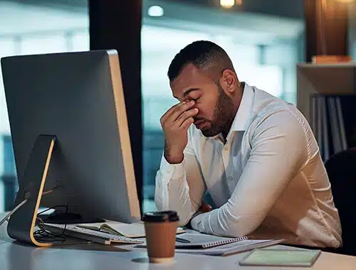 A stressed young African American businessman sits in front of his computer, pinching the crest of his nose with closed eyes to contemplating how to fix his computer issue.