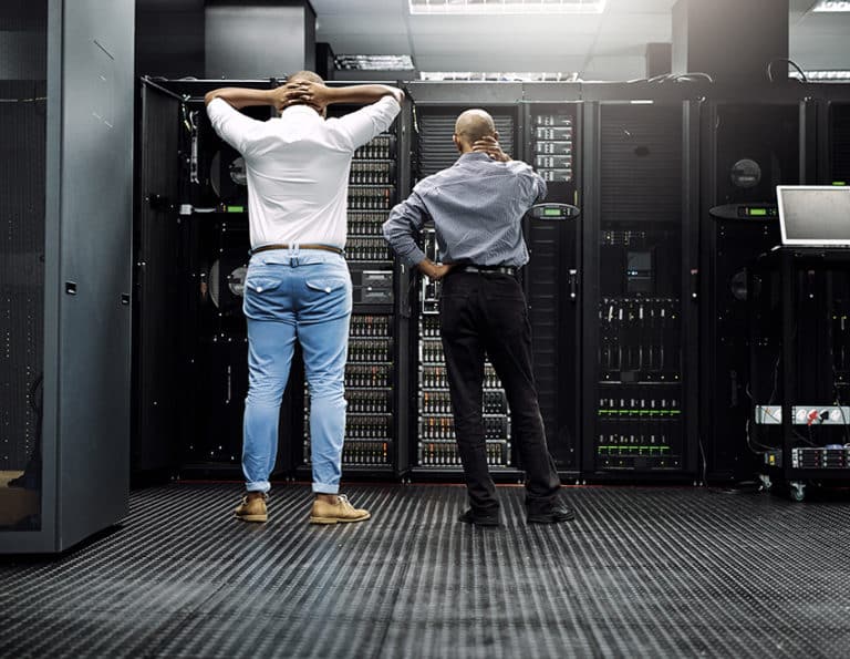 An African American network administrator with his fingers interlocked behind his neck and a system administrator rubbing the back of his kneck appear stressed while looking at a server rack trying to analyze why a server is down and how to restore service quickly for the managed IT services’ business.