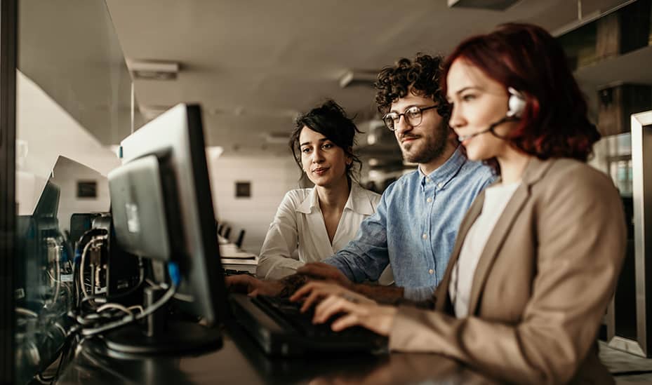 Three help desk professionals are seen working in front of desktops with multiple screens. One of the IT specialists is seen speaking into a headset while resolving a support ticket of a managed services customer.
