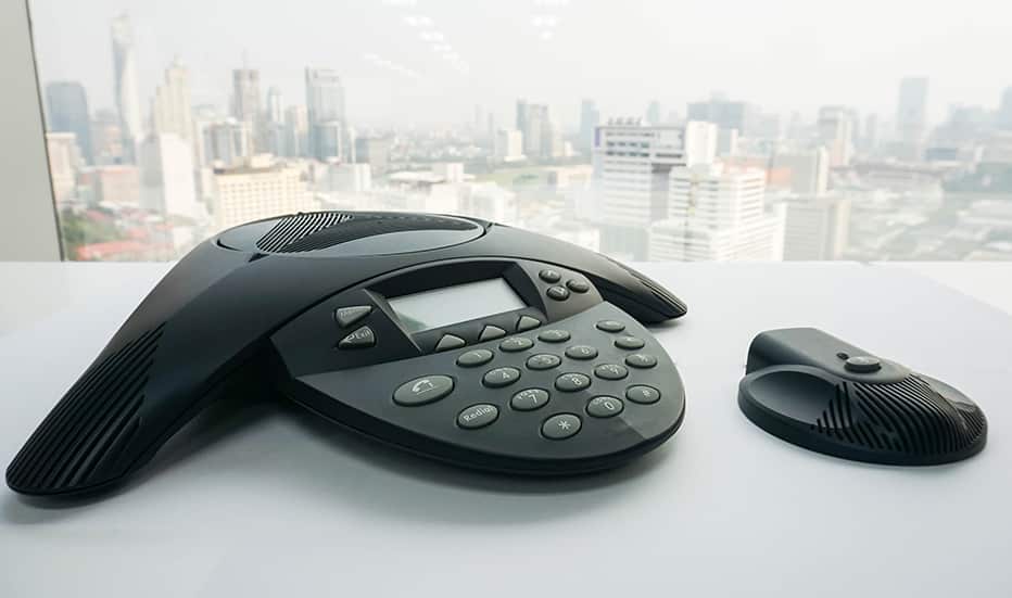 Close up shot of a Polycom VTX 1000 Voice over Internet Protocol business conference phone system sitting on a white desk with a large window in the background overlooking a large city landscape of skyscrapers on a sunny day.