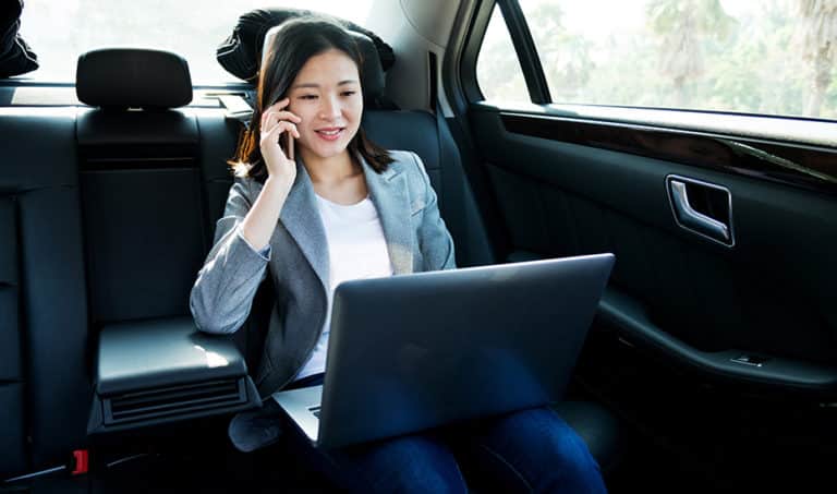 A businesswoman working on her laptop and speaking with a colleague while being chauffeured in a car.