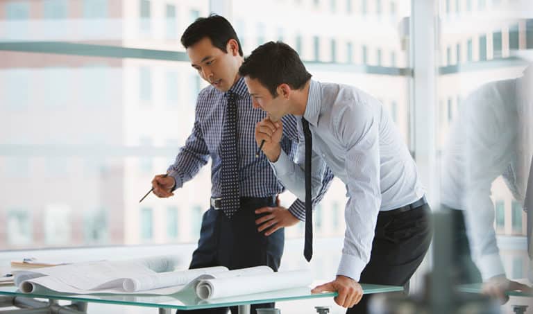 Two middle-aged businessmen collaborate while leaning over a glass desk with scrolls of schematic design plans laid out.