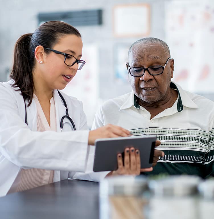 A senior man of African descent meets with his doctor inside a hospital room. The doctor is holding a table and reviewing his medical history, electronic protected health information (ePHI) and discussing treatment options.