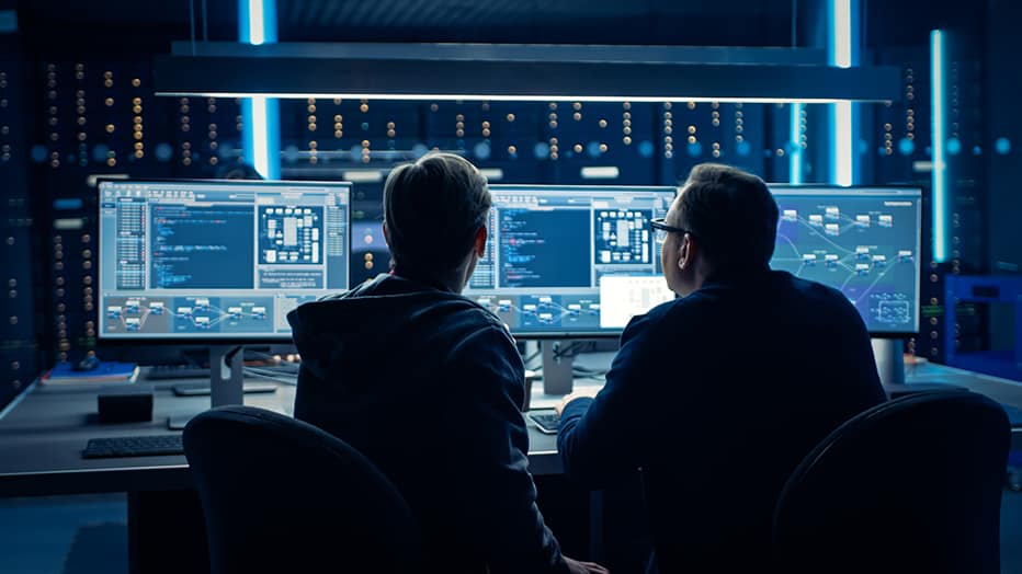 A team of two cybersecurity specialists sitting in front of three monitors, discussing vulnerabilities and the best way to harden the security of a network after a penetration test.