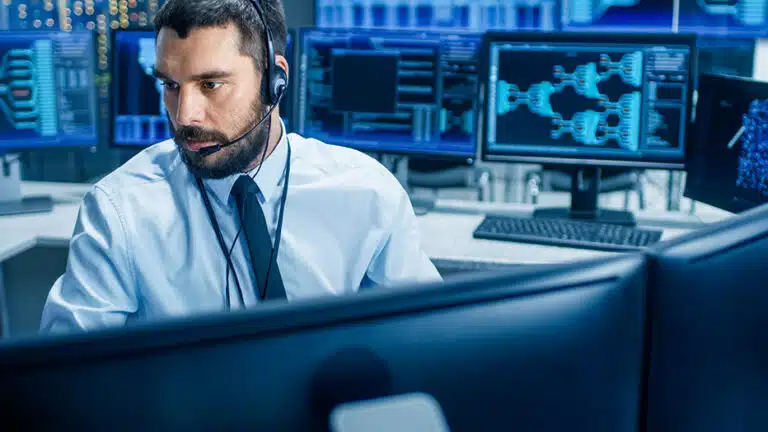 A cybersecurity specialist in a white shirt and black tie wearing a headset monitors several screens for security alerts to detect and rapidly respond to security alerts.