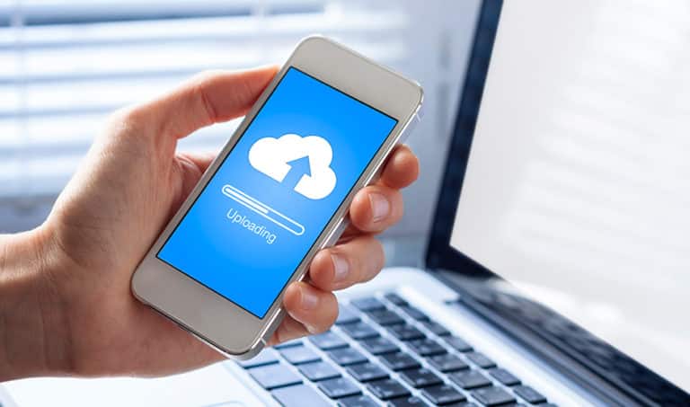 An employee is seen actively backing up important data to the cloud to prevent data loss and ensure business continuity. The mobile phone and laptop show a cloud icon with an arrow pointing up and an uploading progress bar.