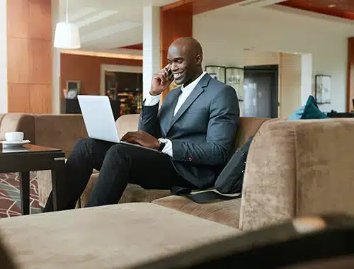 Happy, well-dressed, young businessman sitting on a sofa in a hotel lobby using wifi on his laptop while speaking with a client on his cell phone.