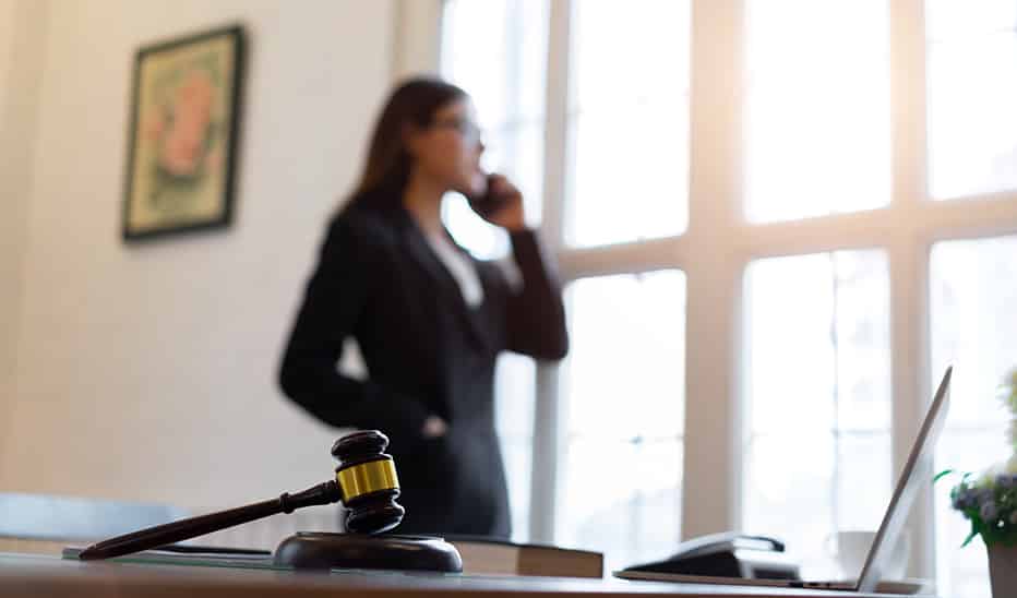 A lawyer consults with a client on her mobile phone while a laptop and gavel are on her desk.