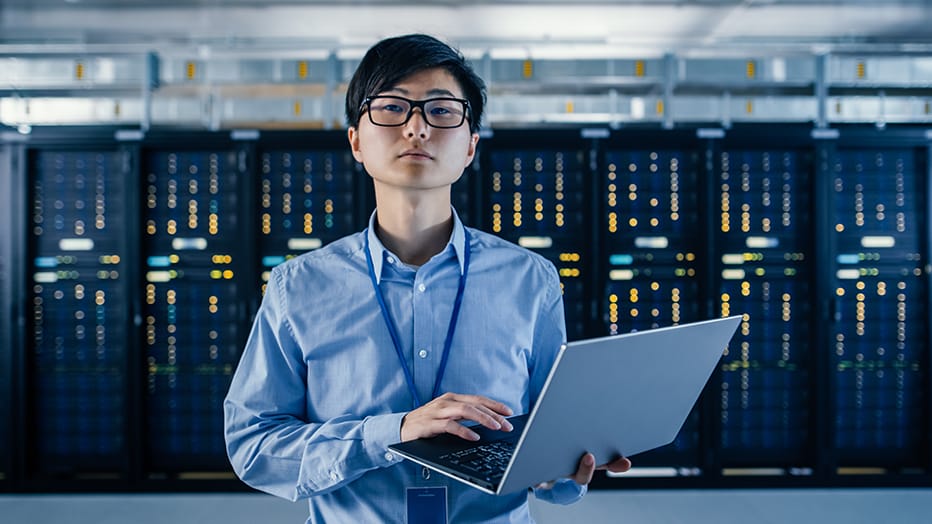 In the modern data center, a network security administrator is standing with server racks behind him, holding a laptop, and looking at the camera with resolute confidence in his ability to defend against a cyberattack.