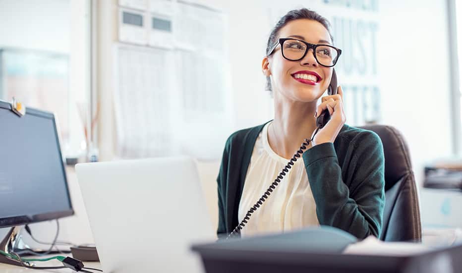 Smiling young businesswoman talking on a managed voip desk phone. Female professional is using a Voice over Internet Protocol business telephone while looking away in a modern office with a large window in the background overlooking the city.