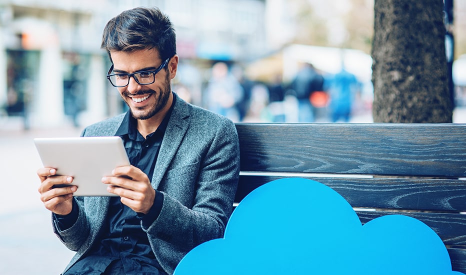 A young business owner accessing documents on-the-go from his iPad sitting on a bench with a blue cloud-cut out used to represent cloud services.