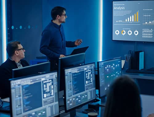 A virtual CIO reviews technology and security strategy presentation with colleagues inside the security operations center, server racks and multiple workstations with monitors can in front of several employees.
